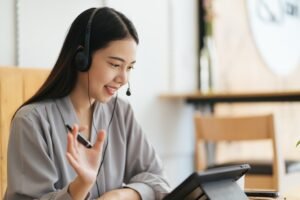 Cheerful beautiful Asian woman wearing headphones and look to digital tablet at video calling meetin